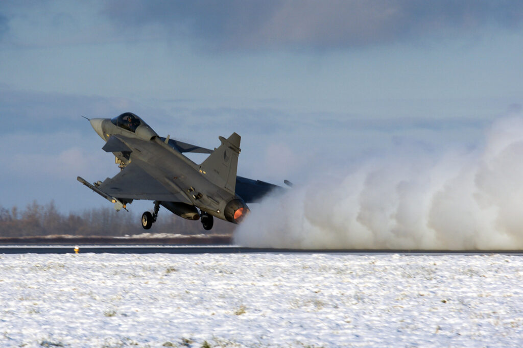 Vojenská stíhačka JAS-39 Gripen vzlétá se zapnutým dohořívačem nad zasněženou ranvejí