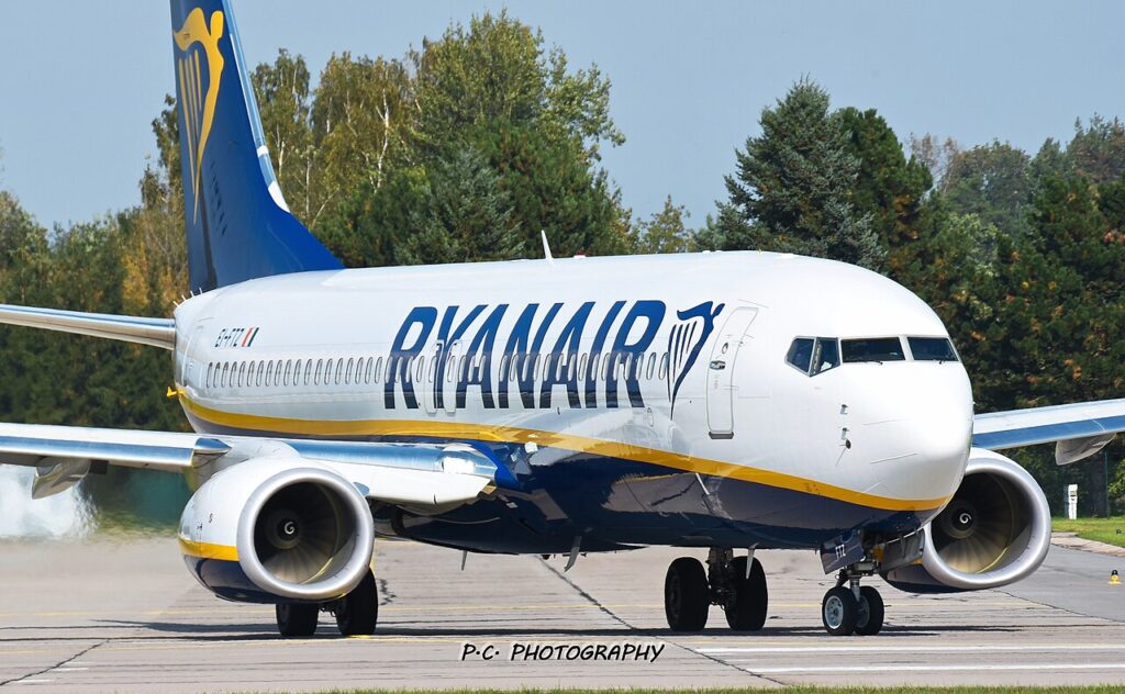 Boeing 737-800 v barvách Ryanairu pojíždí po dráze letiště, v pozadí stromy